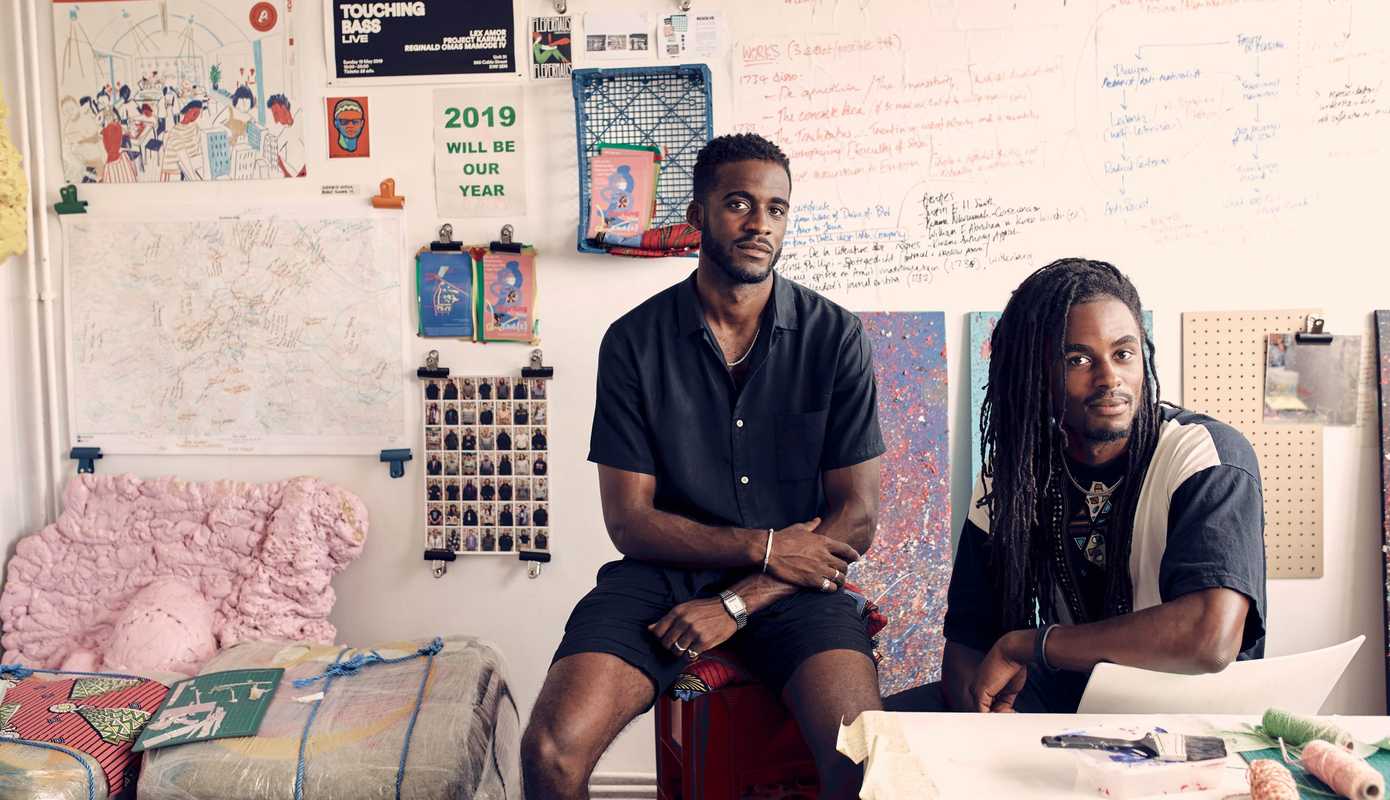 Posed headshot portrait of two men sat down in an art studio, looking straight at the camera with slight smiles