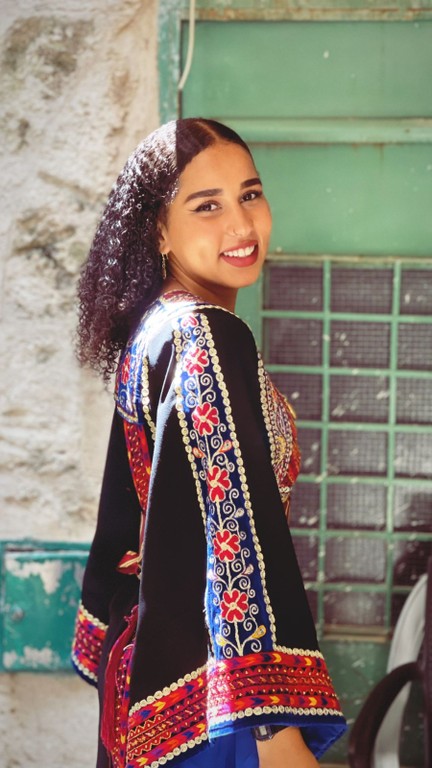 A portrait photograph of a young Afro-Palestinian woman (Shaden Qous). She is turned to the left and smiles at the camera over her right shoulder.