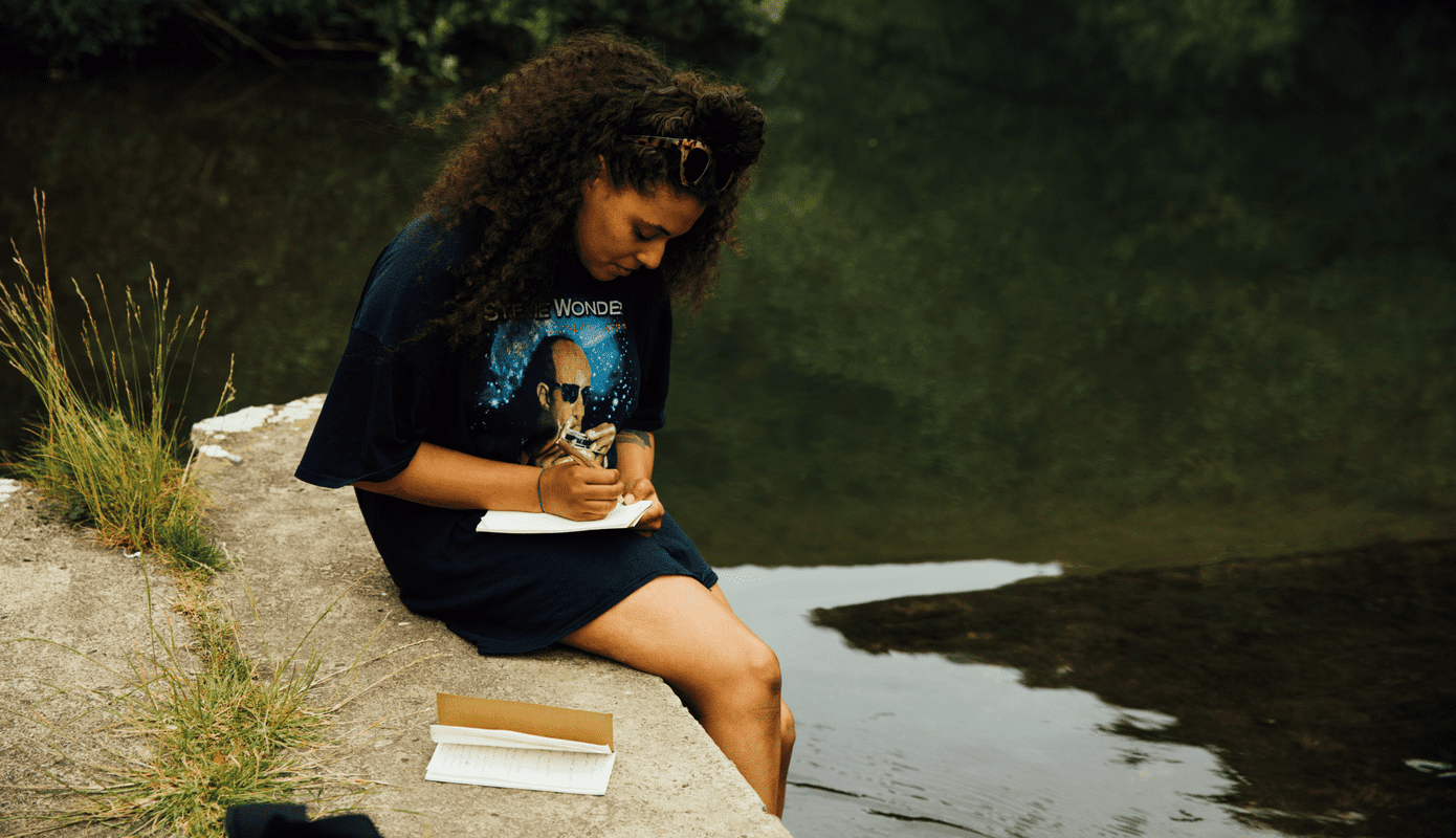 A photograph showing Evie Muir seated on the edge of a river or lake, her feet dangling in the dark water as she is bent over a notebook, pen in hand. She is wearing a t-shirt with Stevie Wonder on it and has sunglasses pushed up onto her head.