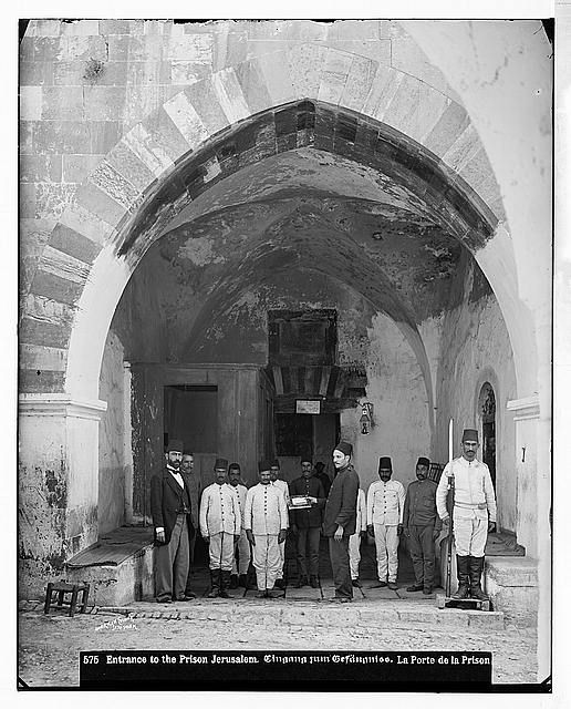 A black and white archival photograph of a group of men standing guard at the arched entrance of a ribat, which was in use as a prison at the time (early 20th century). A caption printed on the photo reads: "Entrance to the Prison Jerusalem"