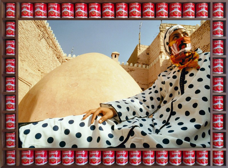 An image of artwork 'Dotted Peace' by Hassan Hajjaj. A photograph of a seated woman wearing a long white garment with black polka dots, a matching head covering and an orange scarf over part of her face. She is in an outdoor setting with architecture visible behind her. Surrounding the photograph are tins of food set into a wooden frame.