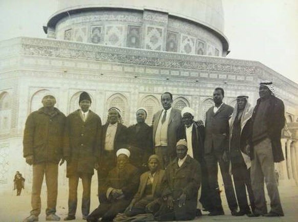 A group of Afro-Palestinian men stand and kneel in a row to have their picture taken in front of the Dome of the Rock. They are wearing clothing from the early 20th century. The photograph is black and white, and is turning yellow with age.