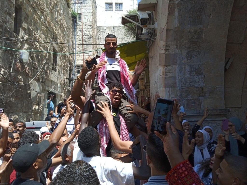 A photograph of Mohammad Firawi, a young Afro-Palestinian man, being carried on the shoulders of a crowd of people celebrating in the narrow alleyways of the African Quarter in Jerusalem.